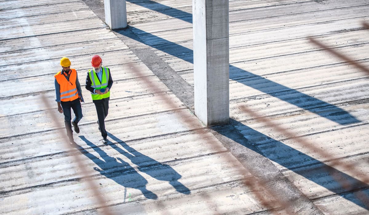 Due tecnici con casco e gilet ad alta visibilità camminano su un solaio in cemento